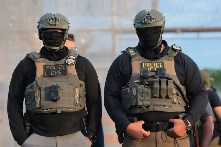 Two masked officers stand in tactical gear marked DHS and ICE.