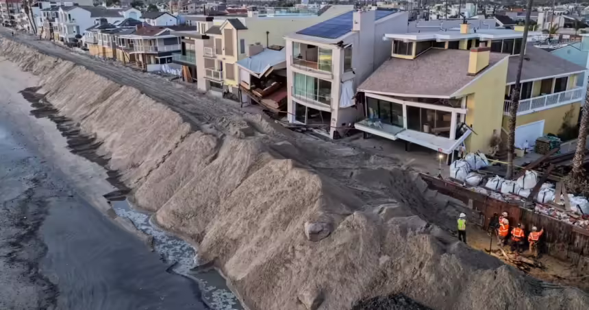 A large sand berm separates beachfront homes from the water as workers stand nearby.